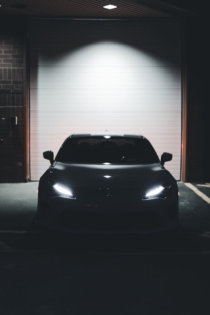 services-01 A dark, mysterious car with glowing headlights parked outside a garage at night.